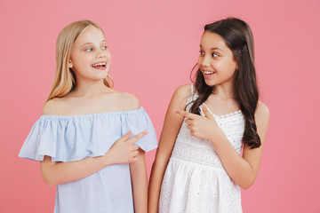Photo of cheerful little girls 8-10 years old wearing dresses smiling and pointing finger at each other, isolated over pink background