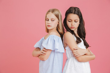 Picture of brunette and blonde girls 8-10 years old wearing dresses standing back to back with arms crossed and expressing quarrel, isolated over pink background