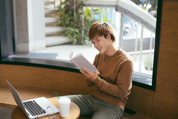 Pensive young asian businessman reading book at desktop with laptop, coffee cup in coffee shop. Knowledge concept