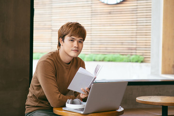 Pensive young asian businessman reading book at desktop with laptop, coffee cup in coffee shop. Knowledge concept