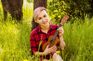 Young handsome girl sitting in green grass and playing with ukulele on wood and bush background
