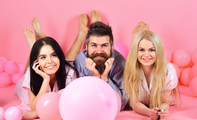 Obraz na płótnie Canvas Blonde and brunette on smiling faces have fun with bearded macho. Relations concept. Best friends, lovers near balloons, pink background. Lovers or best friends in pajamas at girlish bedroom party.