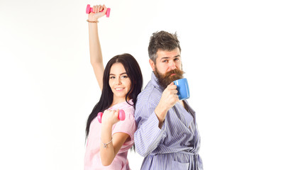 Couple in love in pajama, bathrobe stand isolated on white background. Morning alternative concept. Couple, family on sleepy faces, full of energy. Girl with dumbbell, man with coffee cup.