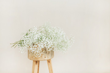 White gypsophila flowers bouquet on wooden backless stool at pale pastel beige background. Minimal...