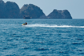 Men in lifejacket riding a white and yellow water scooter on the blue sea, forming large waves, splashes and foam with rocks and ship on the background. 