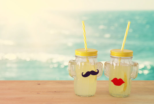 Image of fresh lemonade drink in cute cactus shape glasses wearing mustache and lips, over wooden table. Tropical summer romantic vacation concept.
