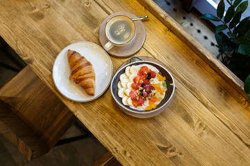  A healthy and tasty breakfast in a cafe of cereal with fruit, a croissant and coffee by the window on a wooden table