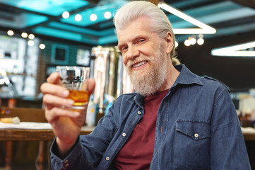 Watching you. Positive delighted senior man raising hand with glass while posing on camera