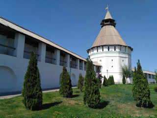 The medieval white stone wall with the loggia and the white stone square tower with the wooden roof and thujas on the sunny day in Astrakhan.