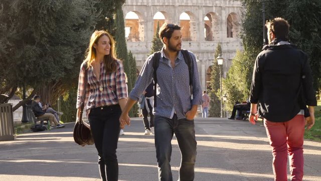 Young happy couple walk in park road with trees and attractive young man glance girlfriend gelousy colosseum in background in rome at sunset lovely beautiful girl with long hair holding hands slow