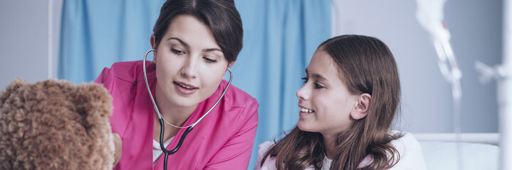Doctor in pink scrubs pretending to listen to the heartbeat with stethoscope, making her young...