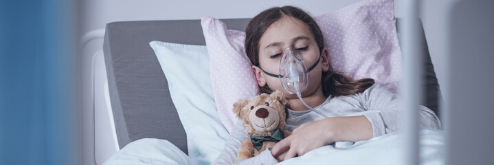 Sick young girl in a hospital bed sleeping with an oxygen mask and a toy, getting treatment for...