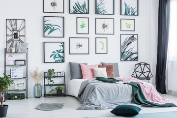 Gallery with simple posters hanging on the wall in bright bedroom interior with many pillows on bed, fresh plants and plastic chair standing next to window with drapes