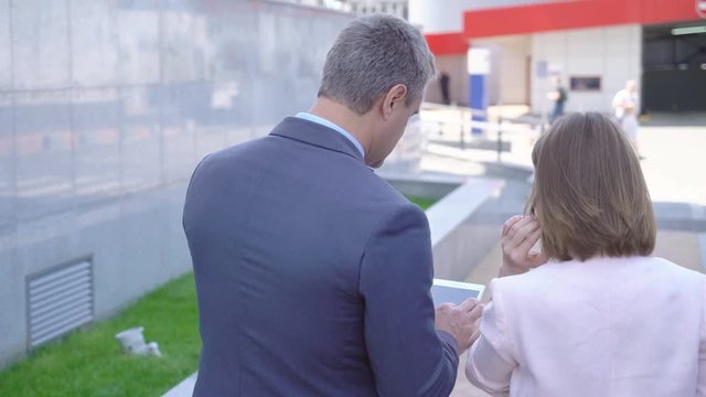 man and woman businessmen walking down the street and discussing a project .. people on the street in the background of an office building are looking at the tablet. business clothes on actors