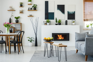 Black chairs at dining table with flowers in flat interior with grey sofa next to fireplace. Real photo