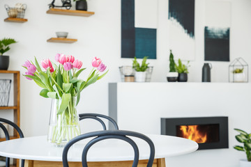 Pink tulips on dining table in white room interior with plants and poster above fireplace. Real photo