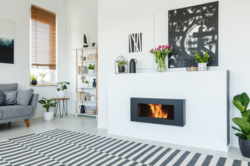 Black painting above fireplace in white living room interior with patterned carpet and grey sofa....
