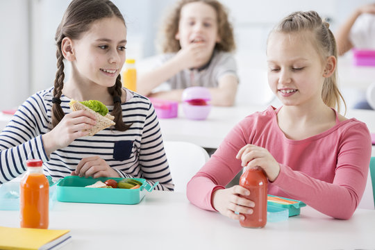 Front view of two junior girls in a school cafeteria during lunch break. One holding a wholewheat sandwich, the other opening a bottle of organic vegetable juice. Other kids in the background