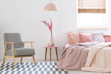 Wooden armchair on patterned carpet in pink bedroom interior with flowers next to bed. Real photo - obrazy, fototapety, plakaty