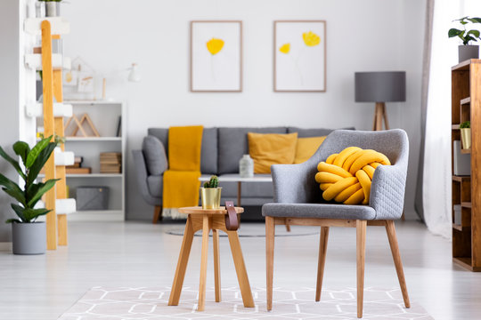Yellow knot cushion on a gray armchair standing next to a wooden stool with a cactus on it in cozy living room interior. Real photo with blurred background