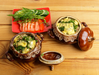 Potatoes are baked in a pot of meat and cheese and a plate of cucumbers and tomatoes on a wooden table.