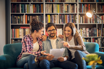 A young handsome male student is surrounded by two beautiful female friends as they go through thesis they worked on together while sitting on a sofa in a library.