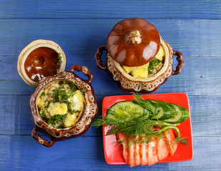 Potatoes are baked in a pot of meat and cheese and a plate of tomato and cucumber slices on a blue wooden background.