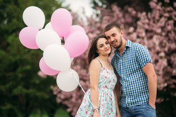 Young happy couple in love outdoors. Handsome man and beautiful woman on a walk in a spring blooming park. They keep helium balls