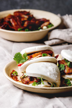 Asian sandwich steamed gua bao buns with pork belly, greens and vegetables served in ceramic plate on table with linen tablecloth. Asian style fast food dinner.