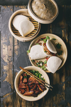 Asian sandwich steamed gua bao buns with pork belly, greens and vegetables served in ceramic plate over dark wooden plank background. Asian style fast food dinner. Flat lay, space