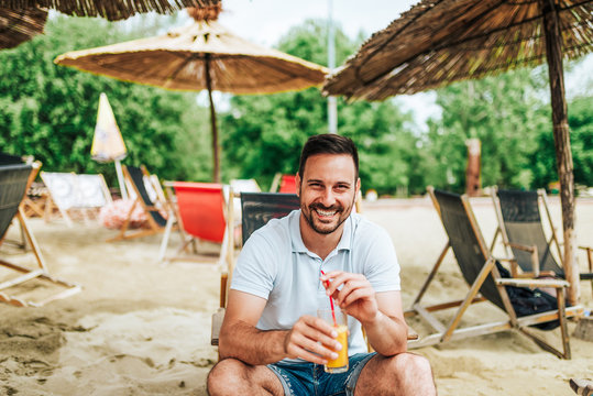 Young Smiling Man Sitting On A Beach Chair And Holding A Cocktail. Looking At Camera.