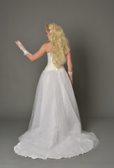Fototapeta premium full length portrait of blonde girl wearing white gown, standing pose with back to the camera. grey studio background.