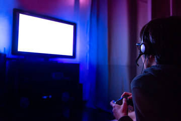 Obraz na płótnie Canvas Young male gamer with glasses and headset playing video game at home in the dark room using game console controller watching at LED TV. Gaming and entertainment concepts