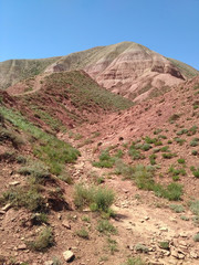 The mountain with red scree slopes covered by green grass. The sky is clear and blue. There is the dried creek bed. It is the sunny summer day.
