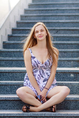 Portrait of a beautiful european woman sitting on steps legs crossed, long blonde hair, sunglasses holds in hands