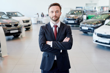 Portrait of handsome sales manager posing confidently with arms crossed standing in car showroom...