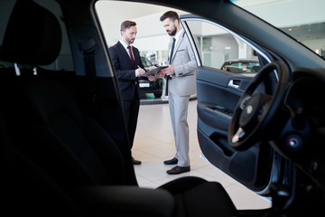 Full length portrait of handsome car salesman talking to customer in showroom, shot from inside...