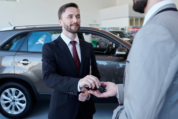 Waist up portrait of smiling handsome car salesman giving car keys to client standing in showroom...