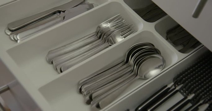 Tilt down to close-up woman's hand as she opens drawer full of stainless steel cutlery, then closes it again in kitchen. Slow motion 4K recorded at 48fps