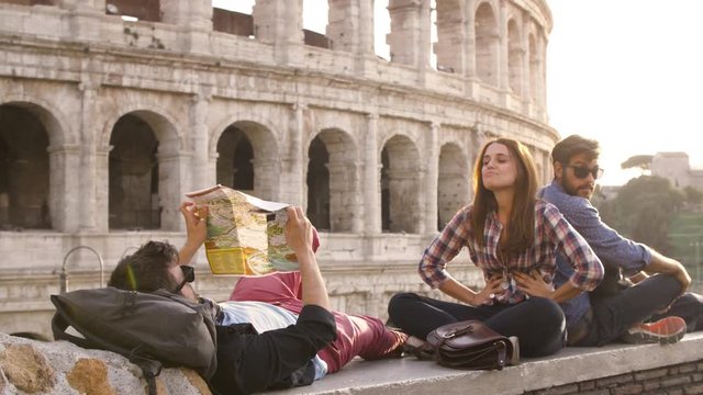 Three young friends tourists sitting lying in front of colosseum in rome at sunset reading map guide for directions pointing with backpacks sunglasses happy beautiful girl long hair slow motion