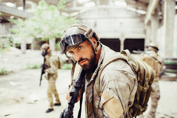 Serious and concentrated warrior is standing and looking straight forward. He is black rifle in his hands. Other two men are looking around and checking the building.