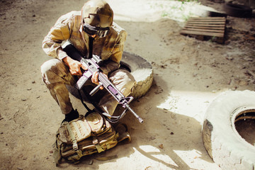 Fototapeta premium A picture of soldier sitting on the ground and wearing face mask. He is holding black rifle in hands. Man is looking odwn to bag. He has some rest.
