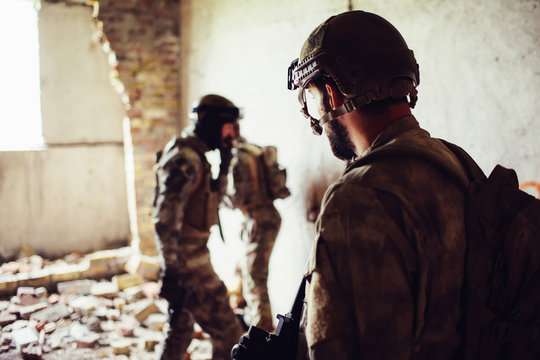 A ppicture of soldiers standing in a rooom near ruined wall. Two of them are standing towards each other while thrid one is looking at them. They are having some rest.