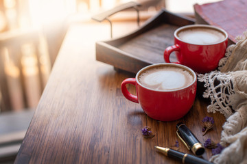 Two red coffee cups on a wooden tray has a scarf, and a pen on the wood table