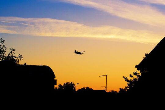 Biplane flying over roofs and trees in vibrant sunset