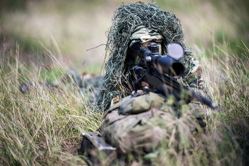 Sniper laying on the grass looking through scope at the target in deep forest.