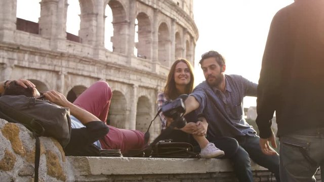 Three young friends tourists sitting lying in front of colosseum in rome at sunset taking pictures photos with dslr camera backpacks sunglasses happy beautiful girl long hair slow motion