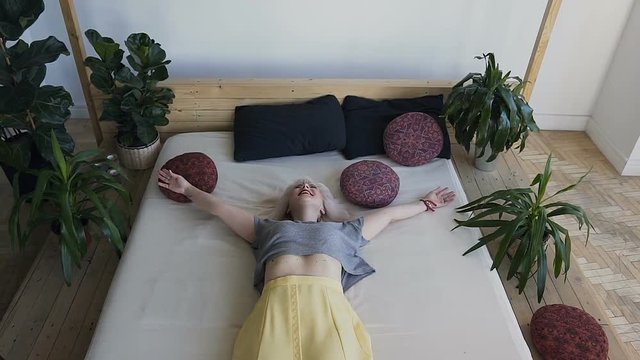 Top view, beautiful blonde fun lies in the big bed and breaks her arms. Smiling girl in a white bed against a background of large green plants. The concept of the morning bedroom of happiness and joy.