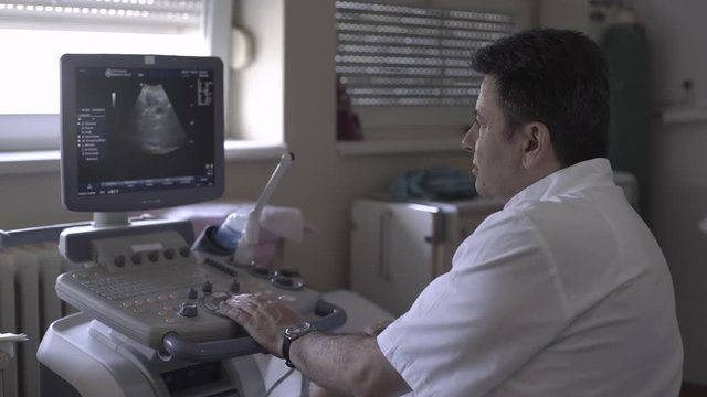 Portrait of male doctor gynecologist while looking at screen and using ultrasound for gynecological examination of woman, female patient lying in ambulance room at gynecology ward, healthcare, indoor
