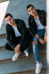 two guys in black jackets, jeans and white shoes are sitting on the steps. on the hands of a watch. socializing, meeting friends.